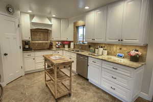 Kitchen with stainless steel dishwasher, premium range hood, white cabinetry, a sink, and stone finish flooring