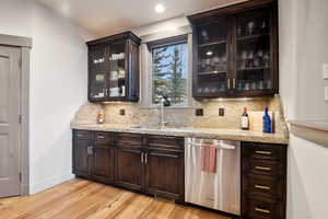 Bar with decorative backsplash, stainless steel dishwasher, light wood-type flooring, a sink, and baseboards
