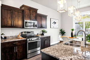 Kitchen featuring appliances with stainless steel finishes, light stone countertops, dark brown cabinets, a sink, and a chandelier