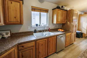 Kitchen with brown cabinetry, dark countertops, dishwasher, a sink, and light wood-style flooring