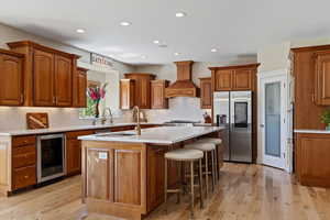 Kitchen featuring stainless steel fridge, beverage cooler, an island with sink, premium range hood, and recessed lighting