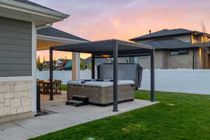 Patio terrace at dusk with outdoor dining area, a hot tub, pergola, lawn, and a fenced backyard