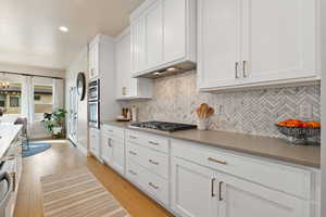 Kitchen featuring white cabinetry, light hardwood flooring, stainless steel gas cooktop and wall oven, a tasteful backsplash and custom range-hood
