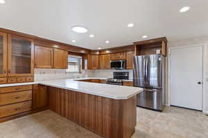 Kitchen featuring a peninsula, glass insert cabinets, light countertops, brown cabinets, and stainless steel appliances