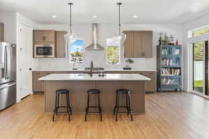 Kitchen with light wood-type flooring, wall chimney exhaust hood, appliances with stainless steel finishes, and tasteful backsplash