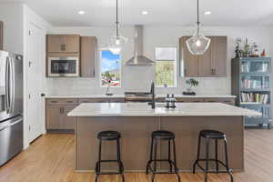 Kitchen with wall chimney range hood, appliances with stainless steel finishes, light wood finished floors, light countertops, and decorative backsplash