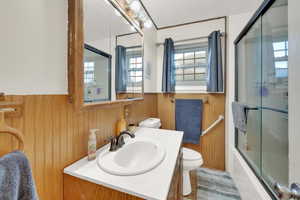 Bathroom with wainscoting, vanity, toilet, wood walls, and bath / shower combo with glass door