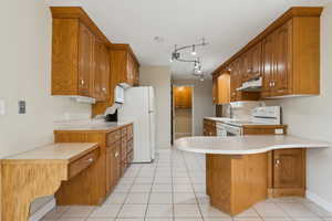 Kitchen with white appliances, brown cabinetry, a peninsula, light tile patterned floors, and under cabinet range hood