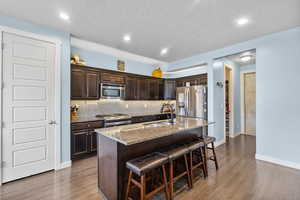 Kitchen with dark brown cabinets, appliances with stainless steel finishes, a sink, wood finished floors, and a kitchen bar
