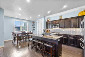 Kitchen with a sink, an island with sink, stainless steel appliances, wood finished floors, and tasteful backsplash
