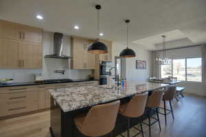 Kitchen with wall chimney range hood, a center island with sink, light wood-type flooring, a sink, and light brown cabinets
