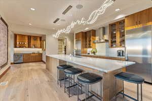 Kitchen with wall chimney exhaust hood, visible vents, beverage cooler, brown cabinets, and high end fridge