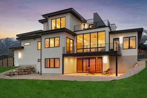 Back of property at dusk featuring a lawn, central AC unit, a patio area, a balcony, and stucco siding