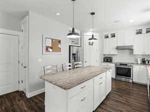 Kitchen with light stone countertops, dark wood-type flooring, under cabinet range hood, appliances with stainless steel finishes, and backsplash