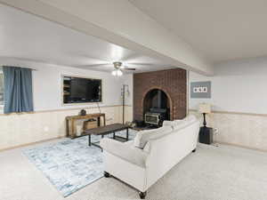 Carpeted living area with wallpapered walls, wainscoting, ceiling fan, and a wood stove