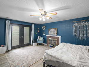 Bedroom featuring ceiling fan, french doors, a textured ceiling, a fireplace with flush hearth, and wood finished floors