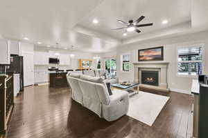Living room with dark wood finished floors, a raised ceiling, ceiling fan with notable chandelier, a warm lit fireplace, and recessed lighting