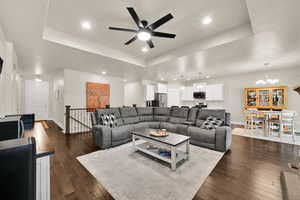 Living area featuring a tray ceiling, dark wood finished floors, baseboards, and ceiling fan with notable chandelier
