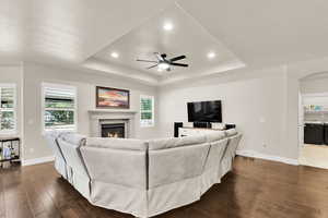 Living area with ceiling fan, a glass covered fireplace, dark wood finished floors, baseboards, and a tray ceiling