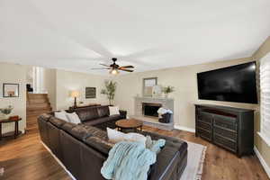 Living area with a ceiling fan, a stone fireplace, wood finished floors, and view of stairs leading to upper level.