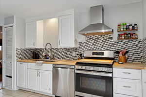 Kitchen featuring butcher block counters, wall chimney exhaust hood, white cabinets, a sink, and appliances with stainless steel finishes