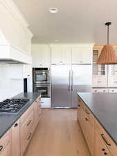 Kitchen with decorative backsplash, decorative light fixtures, appliances with stainless steel finishes, light wood finished floors, and custom range hood