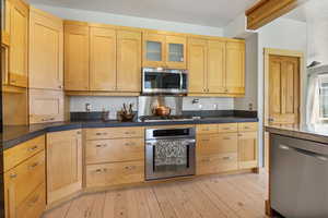 Kitchen with light wood-style floors, glass insert cabinets, light brown cabinets, and stainless steel appliances