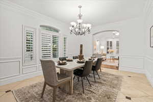 Dining space with crown molding, a decorative wall, arched walkways, a chandelier, and light tile patterned floors