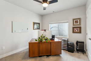 Home office featuring ceiling fan, visible vents, baseboards, radiator heating unit, and light carpet