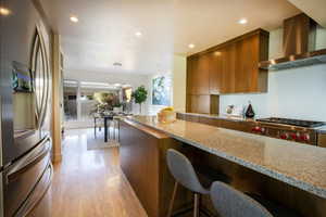Kitchen with brown cabinetry, light wood-style flooring, wall chimney exhaust hood, stainless steel appliances, and modern cabinets
