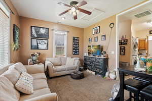 Carpeted living area with stacked washer and dryer, ceiling fan, visible vents, and attic access