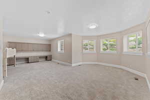Unfurnished living room with baseboards, light colored carpet, built in desk, and visible vents