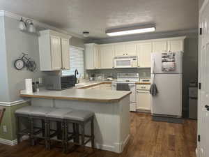 Kitchen with dark wood-style flooring, a peninsula, white appliances, white cabinetry, and a sink