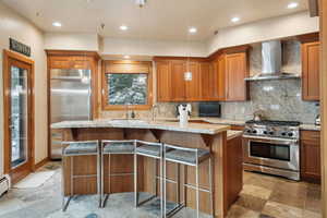 Kitchen featuring wall chimney exhaust hood, light stone countertops, high quality appliances, a sink, and brown cabinets