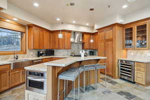 Kitchen featuring light stone countertops, stainless steel oven, wall chimney range hood, a sink, and wine cooler
