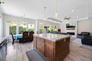 Kitchen featuring brown cabinetry, a fireplace, a raised ceiling, ceiling fan with notable chandelier, and light wood finished floors