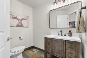 Half bath featuring vanity, visible vents, toilet, baseboards, and stone finish flooring