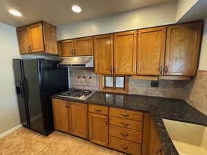 Kitchen with a sink, brown cabinetry, black fridge with ice dispenser, dark stone countertops, and under cabinet range hood