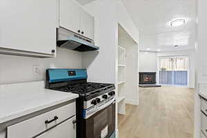 Kitchen with stainless steel gas range oven, under cabinet range hood, a glass covered fireplace, white cabinetry, and light wood-type flooring