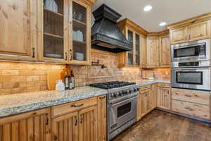Kitchen featuring light stone countertops, custom range hood, decorative backsplash, appliances with stainless steel finishes, and dark wood-style floors