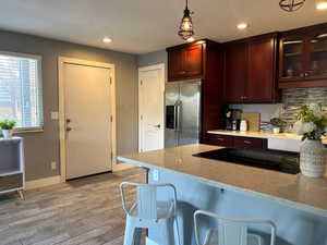 Kitchen with light wood-style flooring, glass insert cabinets, light stone counters, stainless steel fridge with ice dispenser, and black electric cooktop