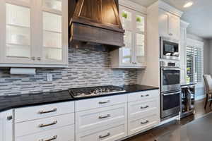 Kitchen with dark countertops, white cabinets, backsplash, appliances with stainless steel finishes, and custom exhaust hood
