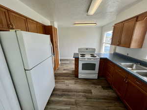 Kitchen with brown cabinetry, white appliances, a textured ceiling, dark wood-style floors, and dark countertops