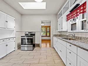 Kitchen with stainless steel range with electric stovetop, a skylight, a sink, white cabinets, and a wainscoted wall