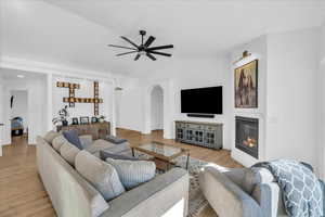 Living room with a ceiling fan, arched walkways, a glass covered fireplace, light wood-style floors, and vaulted ceiling