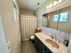 Bathroom with toilet, vanity, curtained shower, visible vents, and a textured wall