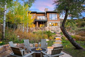 Back of house at dusk featuring a balcony, an outdoor fire pit, stone siding, a patio area, and a chimney