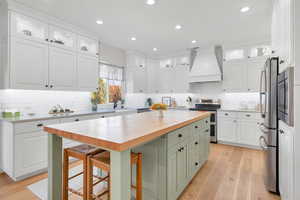 Kitchen with custom exhaust hood, butcher block counters, white cabinets, and appliances with stainless steel finishes