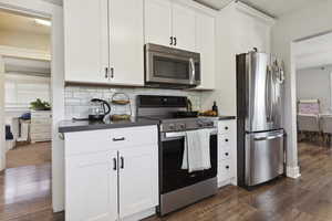 Kitchen with white cabinets, dark wood-type flooring, dark countertops, stainless steel appliances, and tasteful backsplash