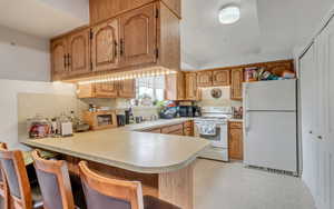Kitchen with brown cabinetry, white appliances, a peninsula, a kitchen breakfast bar, and light countertops